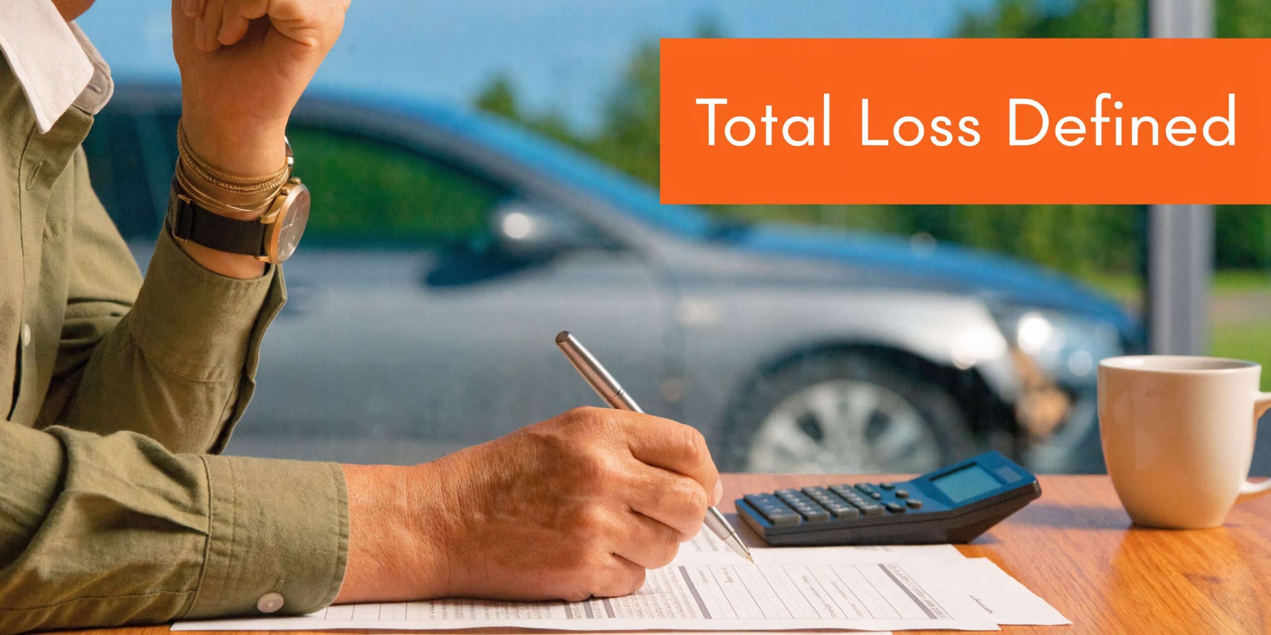 A person writing on insurance documents at a desk with a calculator, coffee, and a damaged car nearby.